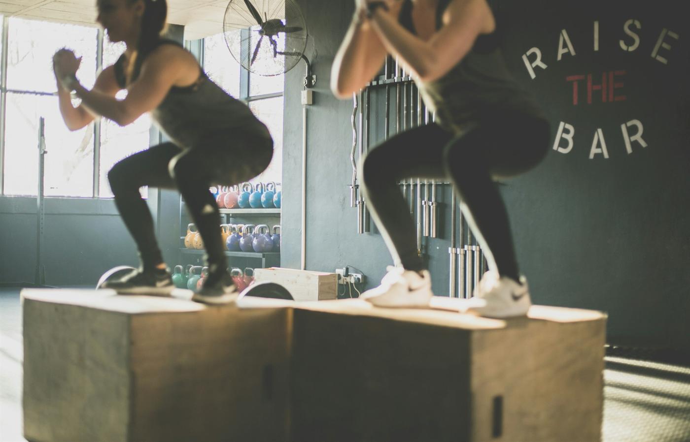 Two athletes mid-jump onto wooden plyo boxes in a bright training studio