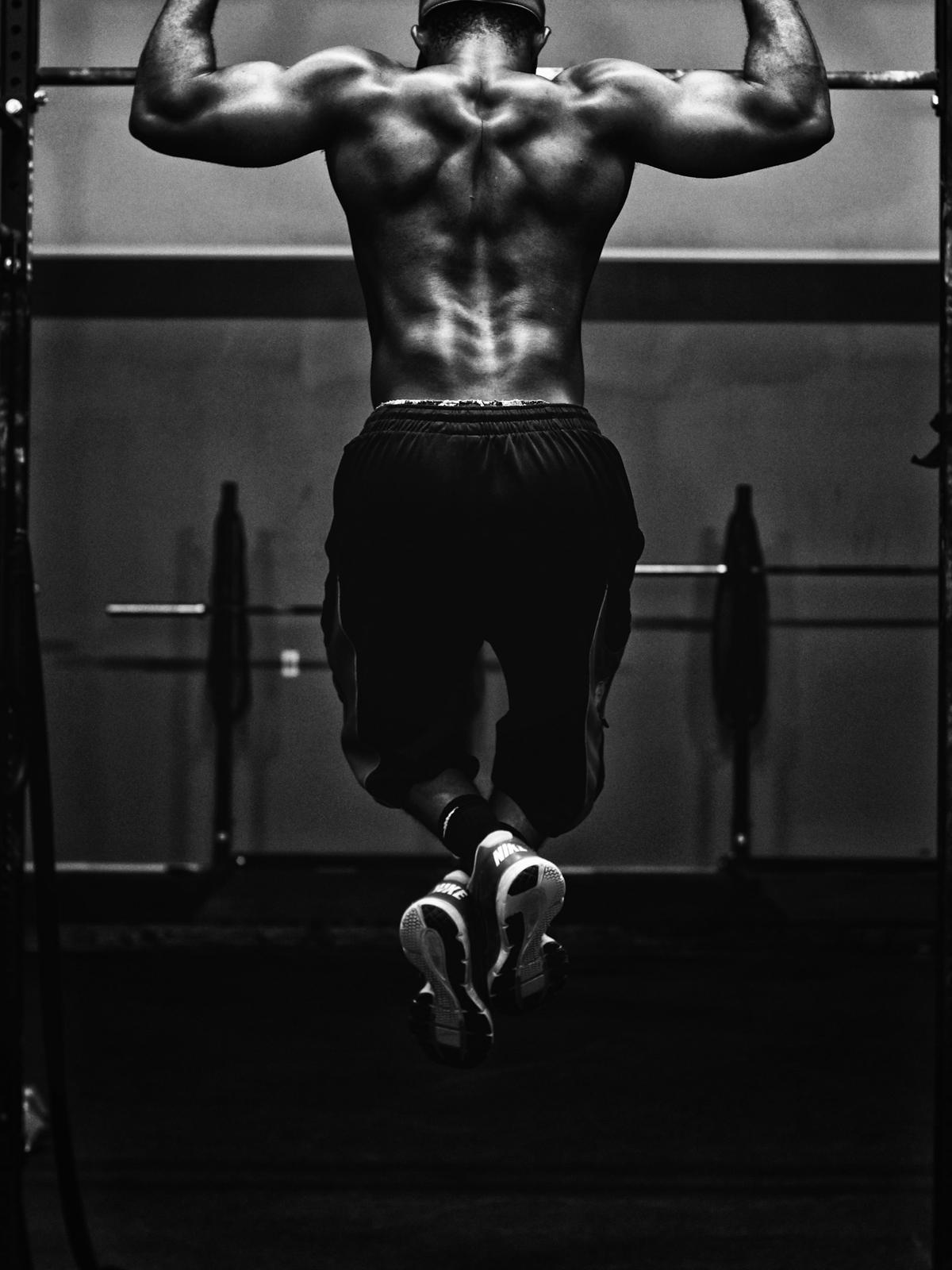 A shirtless athlete seen from behind, hanging from a pull-up bar in a dark gym