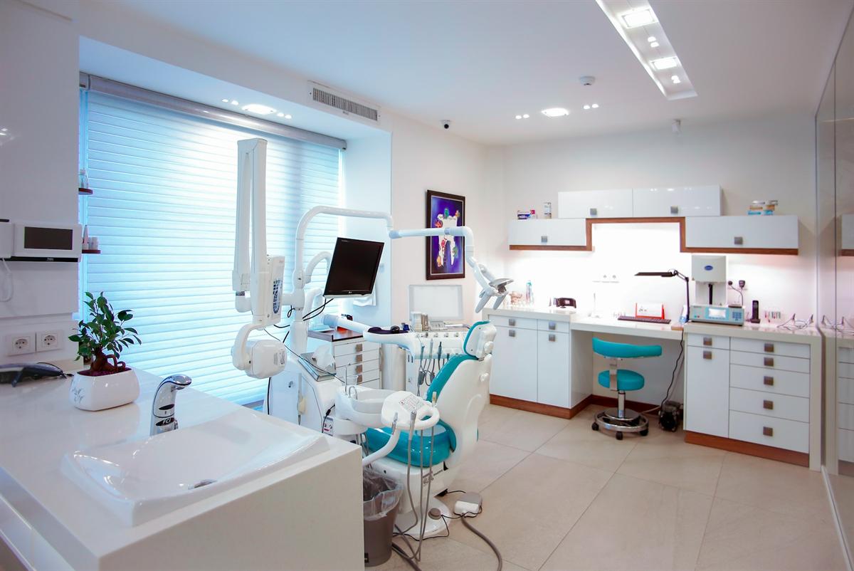 A bright treatment room with a teal dental chair, soft blinds and white cabinetry
