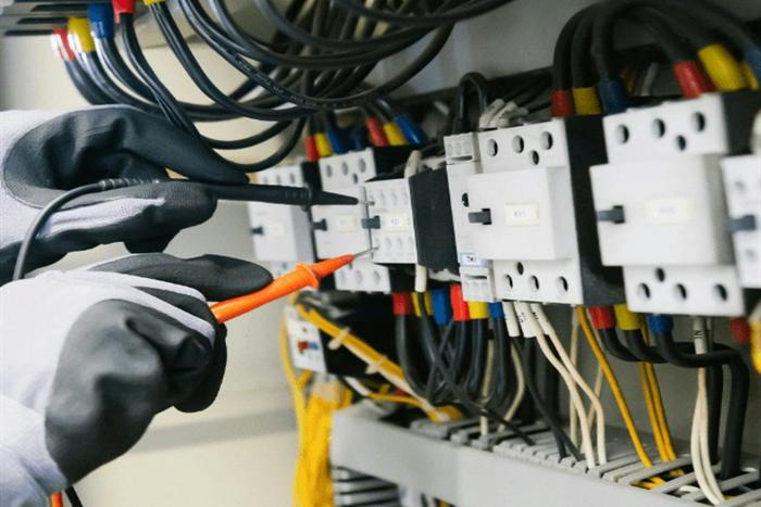 Gloved hands testing terminals inside a distribution board with a multimeter probe